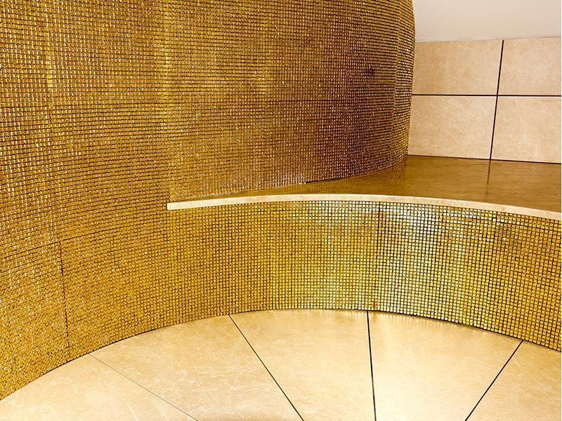 Goldene Mosaikfliesen bedecken eine gebogene Wand und einen Teil des Bodens, im Hintergrund sind beige Fliesen zu sehen.