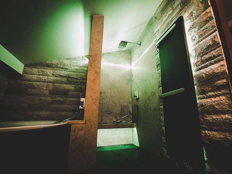 Duschbereich mit Steinwänden und grüner Beleuchtung. Ein Holzbalken und eine dunkle Tür sind zu sehen.