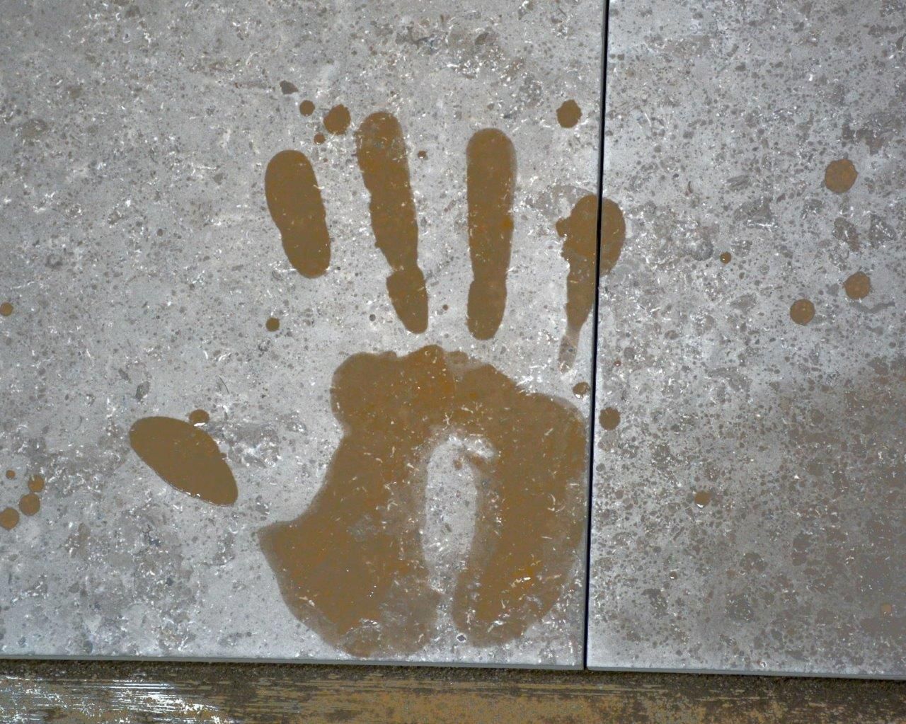 Handabdruck in nasser, schlammiger Substanz auf Steinfliese, mit verstreuten Tropfen.