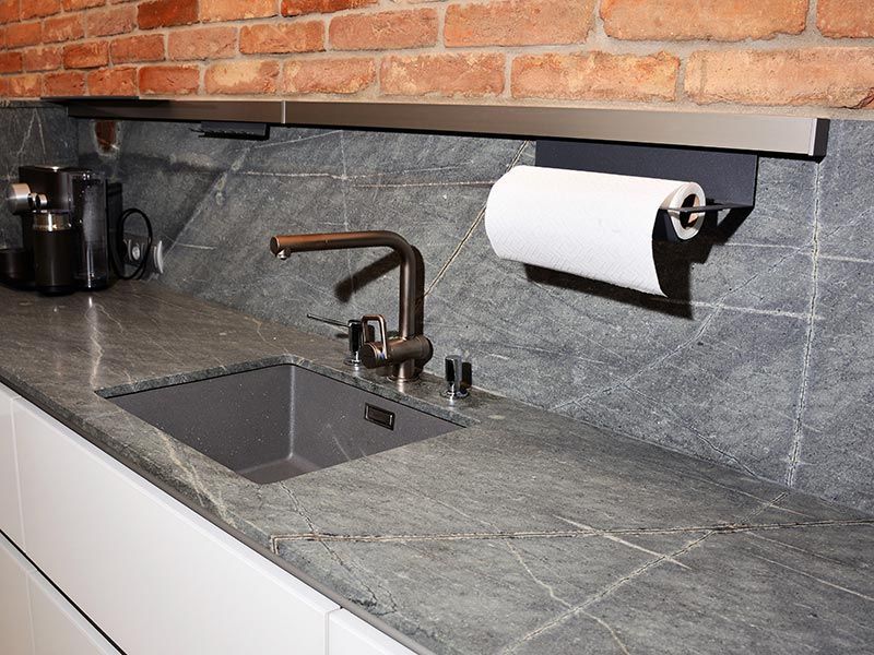 Küchenarbeitsplatte mit Spüle, Wasserhahn und Papierhandtuchhalter; Backsteinwand im Hintergrund.