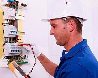 Electricista con camisa azul y casco blanco inspeccionando el cableado de un cuadro eléctrico con un multímetro.