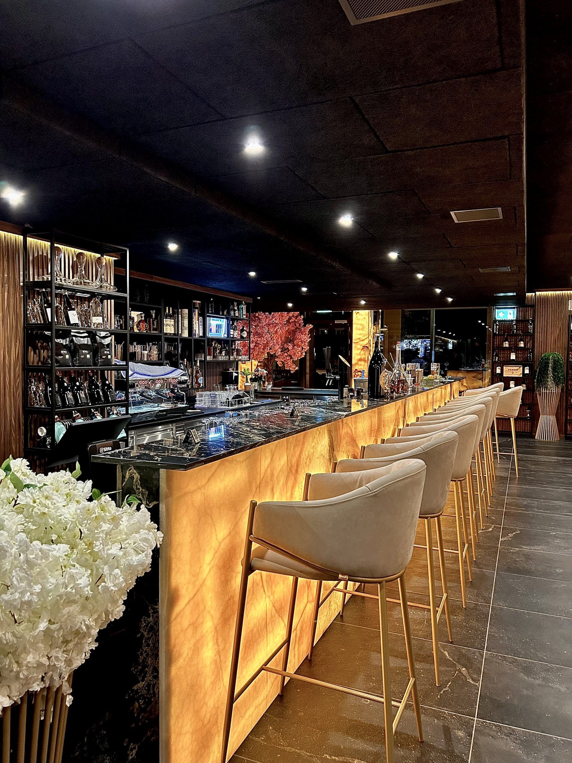 Lange Bar mit goldenen Akzenten und cremefarbenen Barhockern, warm beleuchtet, dahinter sind Flaschen zu sehen.