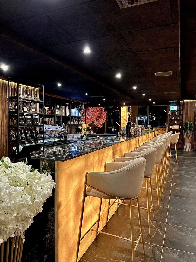 Lange Bar mit goldenen Akzenten und cremefarbenen Barhockern, warm beleuchtet, dahinter sind Flaschen zu sehen.