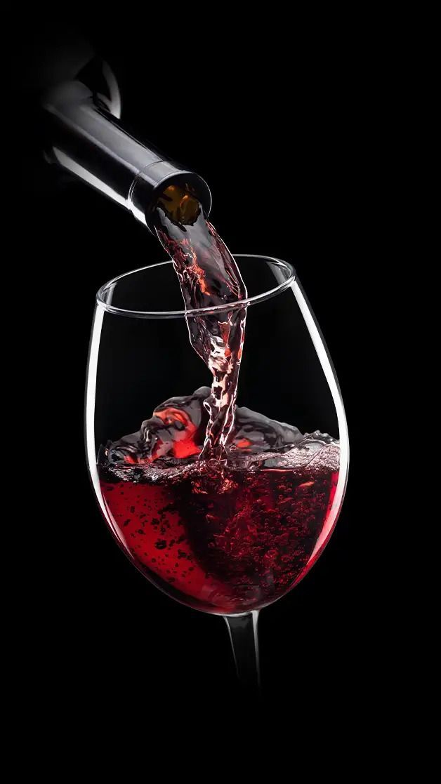 Rotwein wird in ein Glas gegossen, im Hintergrund steht eine Flasche, alles vor einem schwarzen Hintergrund.