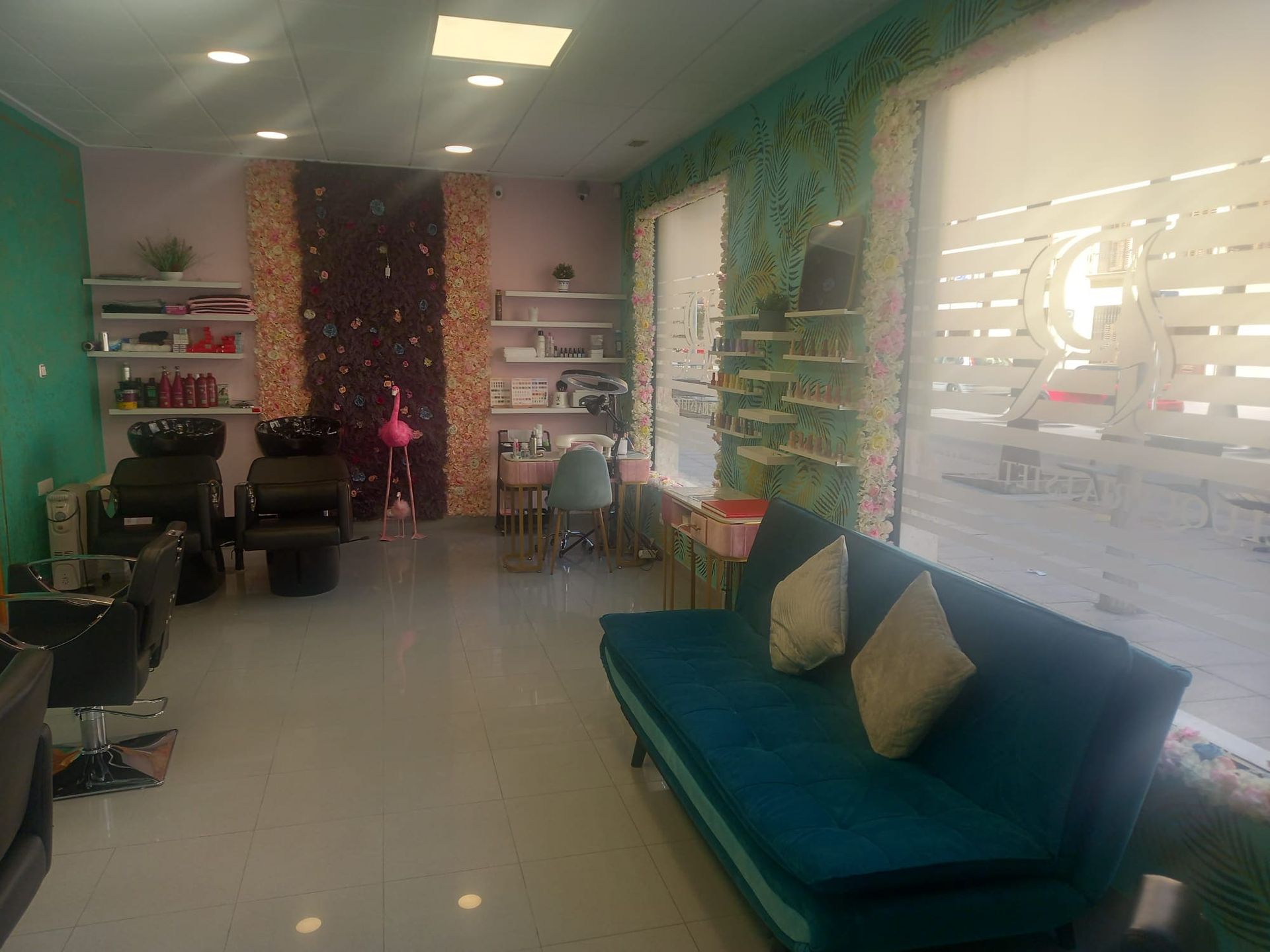 Un sofá de color verde azulado ocupa un lugar destacado en una luminosa peluquería con estaciones de trabajo, estanterías y una pared decorativa con un estampado.