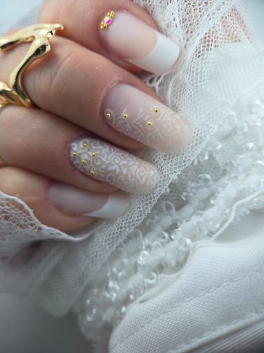 Mano con uñas de estilo nupcial: puntas francesas y diseños de encaje adornados con pequeñas cuentas doradas, sobre un fondo de encaje blanco.