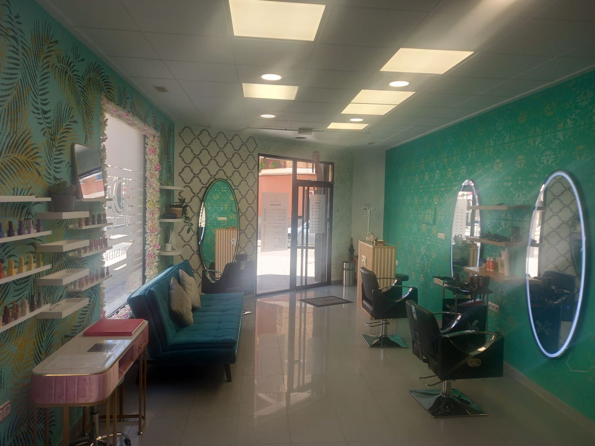 Una peluquería moderna con paredes de color verde azulado, un sofá azul, sillones de peluquería con espejos ovalados y estantes montados en la pared para los productos.