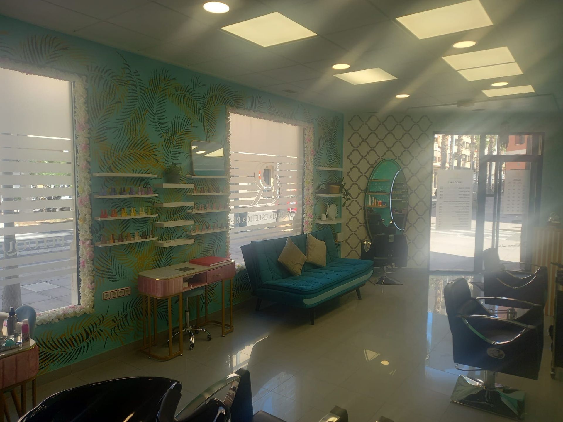 Una peluquería muy iluminada con paredes de color turquesa decoradas con motivos de hojas de palmera, un sofá de terciopelo, sillones de peluquería y estanterías con productos.
