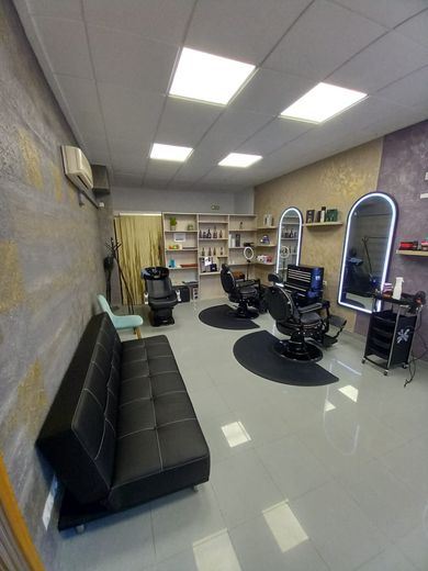 Una peluquería con un sofá negro, dos sillones de peluquería con espejos, una estación de lavado de cabello y estantes con productos.