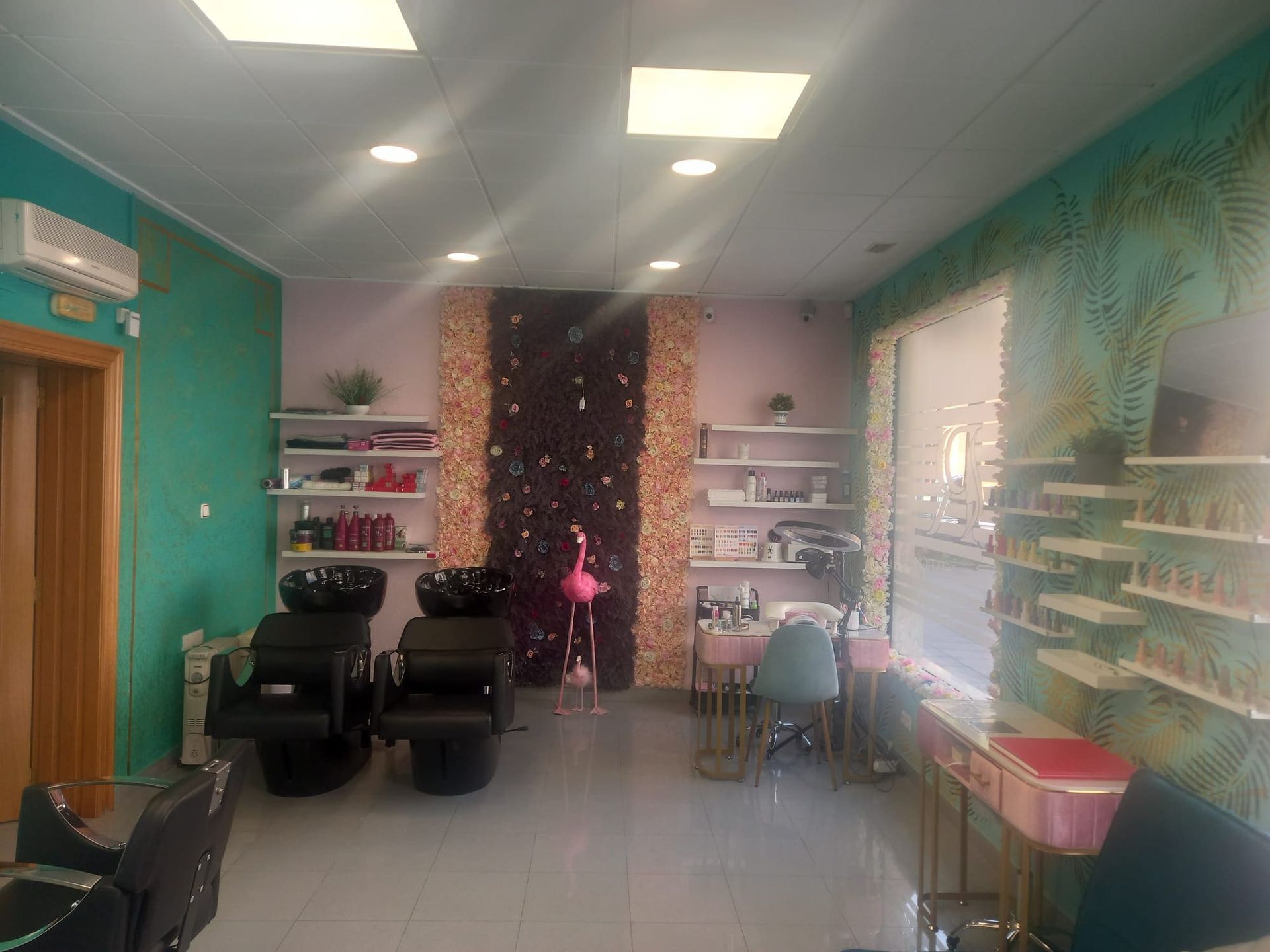 El interior de un salón de belleza cuenta con dos estaciones de lavado de cabello negras, una pared decorativa con motivos florales, decoración con flamencos y estantes para productos montados en la pared.