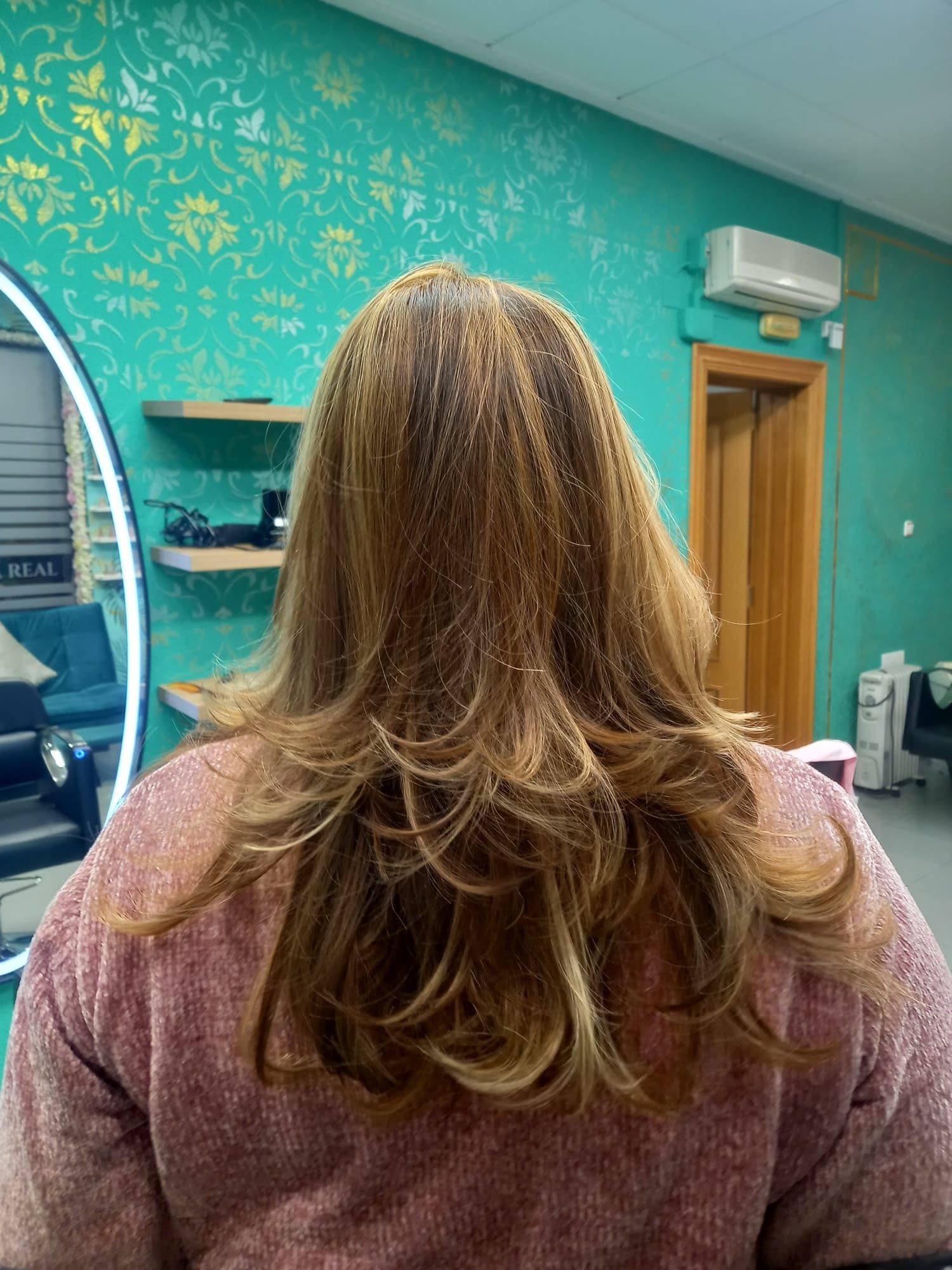 Vista posterior de una melena castaña en capas con reflejos rubios en una peluquería de color turquesa.