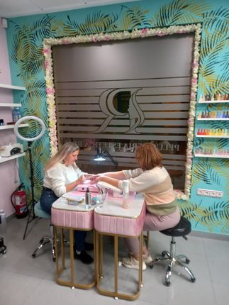 Una técnica de uñas trabaja en las manos de una clienta en una mesa blanca en un salón con paredes decoradas con motivos de palmeras y flores ornamentales.