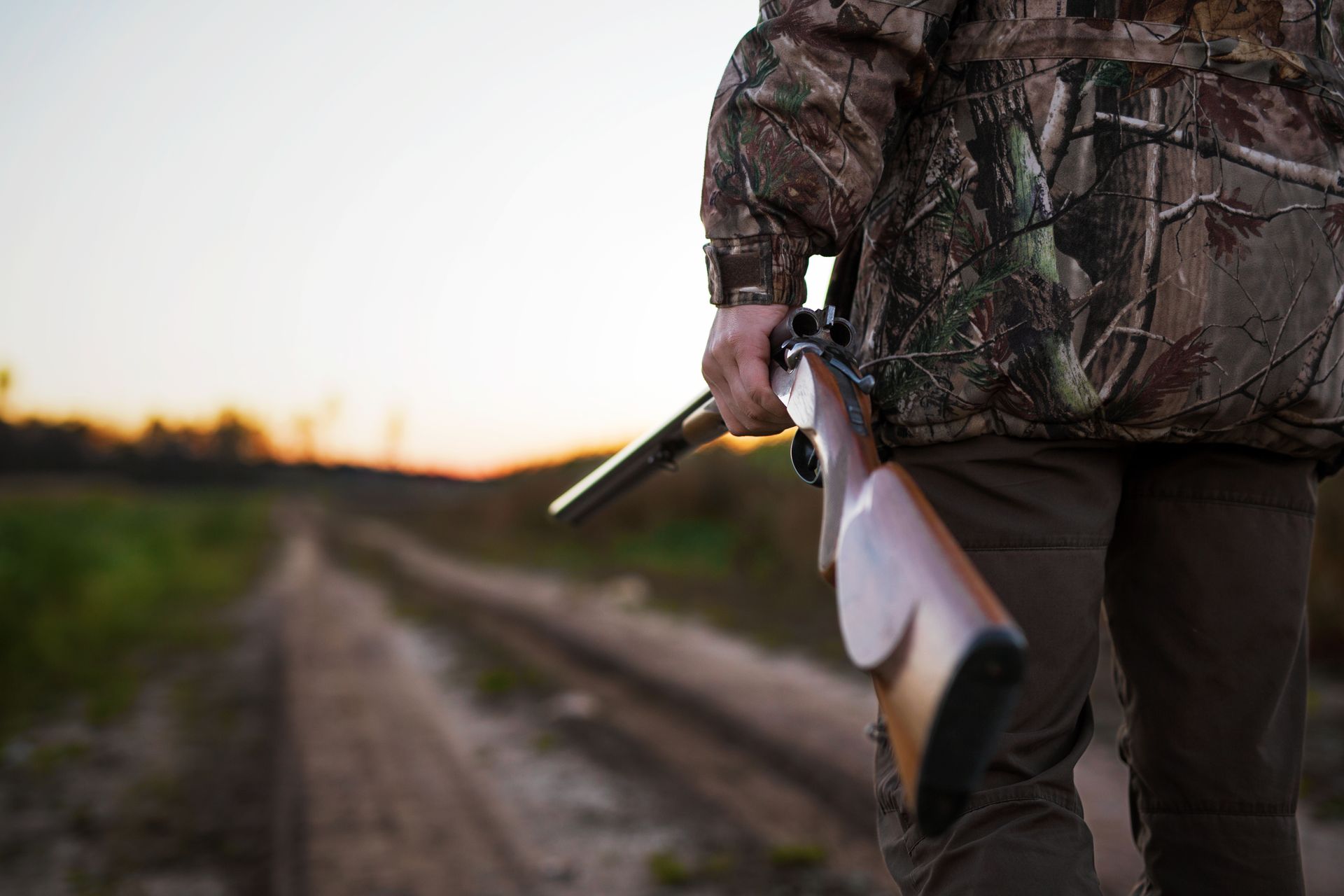 Un chasseur en tenue de camouflage, fusil à la main, descend un chemin de terre au coucher du soleil.