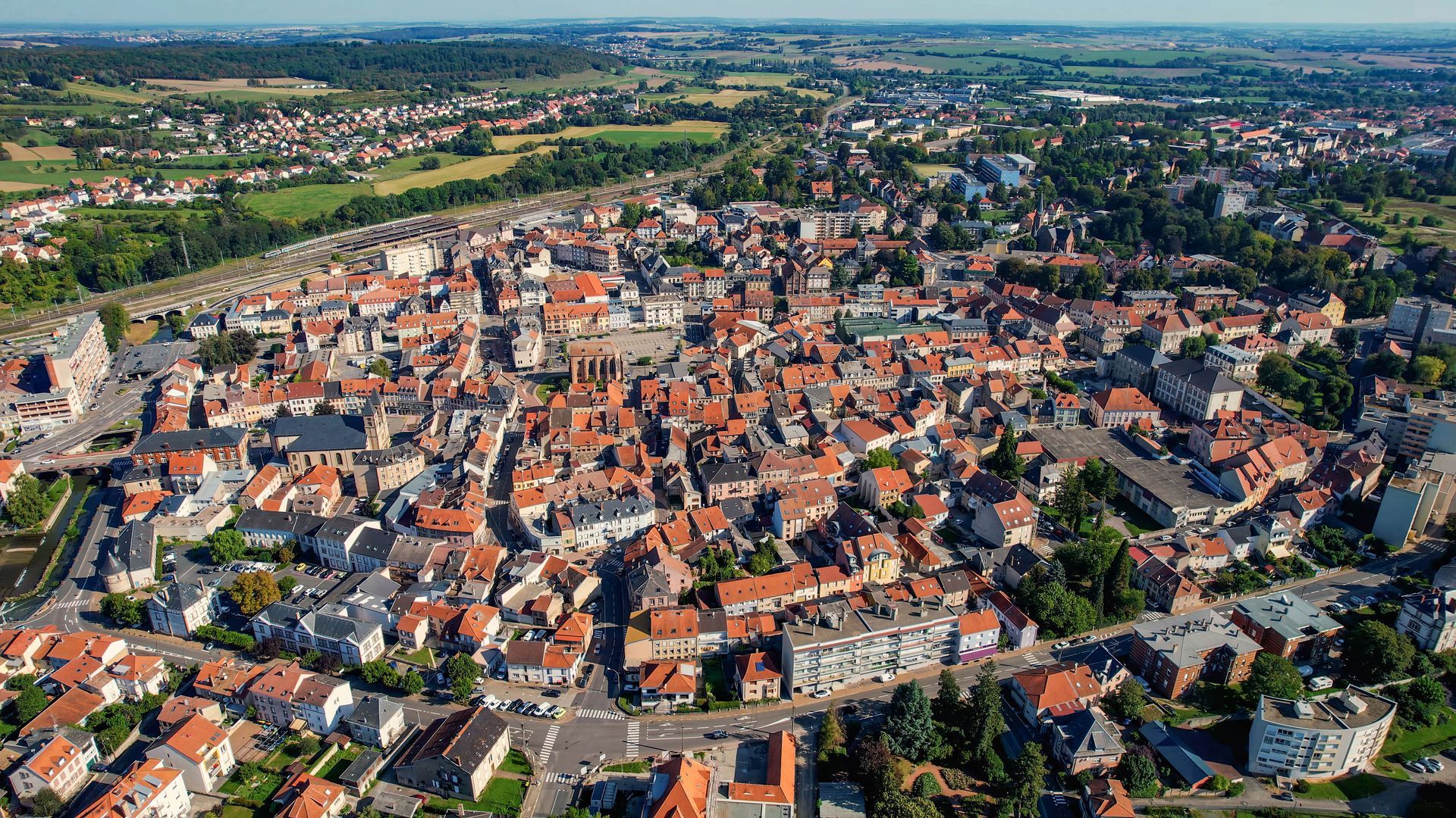 Vue aérienne d'une ville aux toits de tuiles rouges, avec des bâtiments, des arbres et des routes.