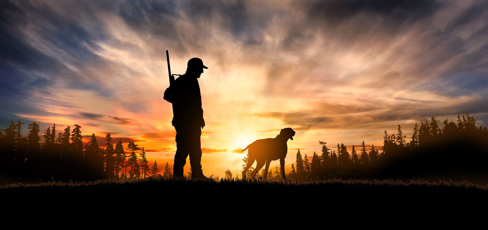 Silhouette d'un chasseur et de son chien dans un champ au coucher du soleil. Le chasseur porte un fusil, le chien est debout. Ciel orange et bleu.
