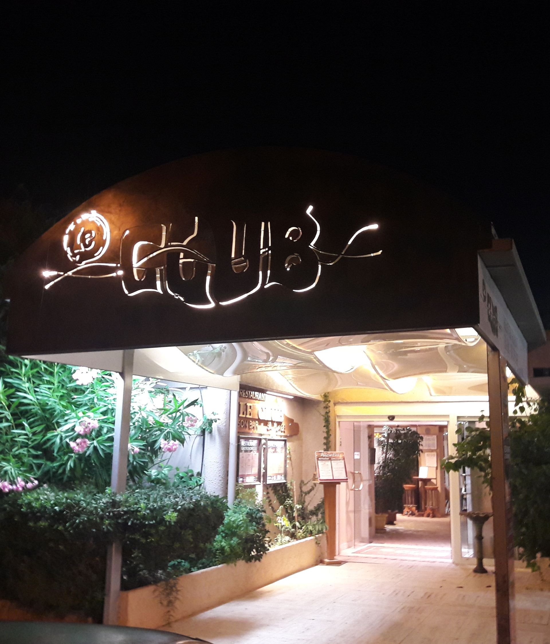 Entrée illuminée d'un restaurant la nuit, avec une enseigne lumineuse et des plantes le long de l'allée.