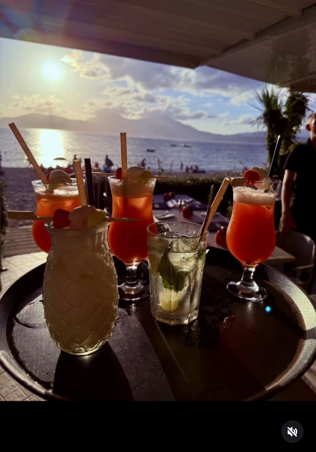 Cocktails sur une table de plage au coucher du soleil, avec l'océan et les montagnes en arrière-plan.