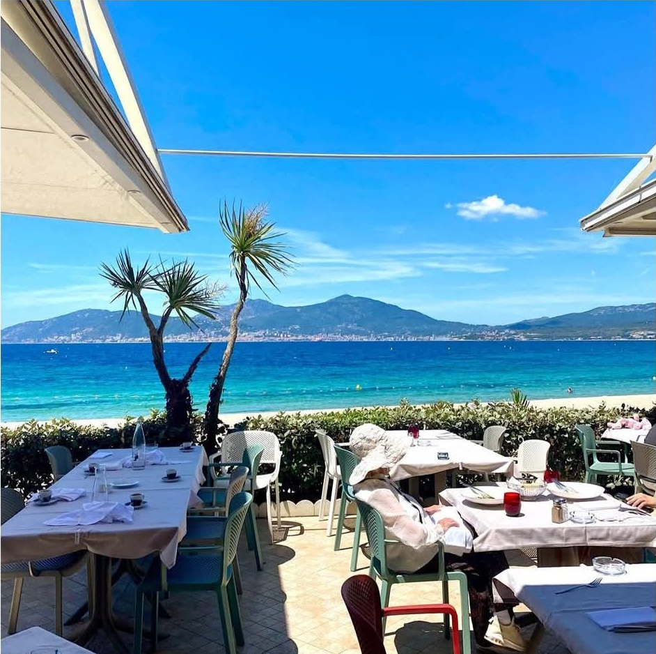 Terrasse de restaurant en bord de mer avec tables blanches et parasols donnant sur un océan d'un bleu éclatant et des montagnes au loin.