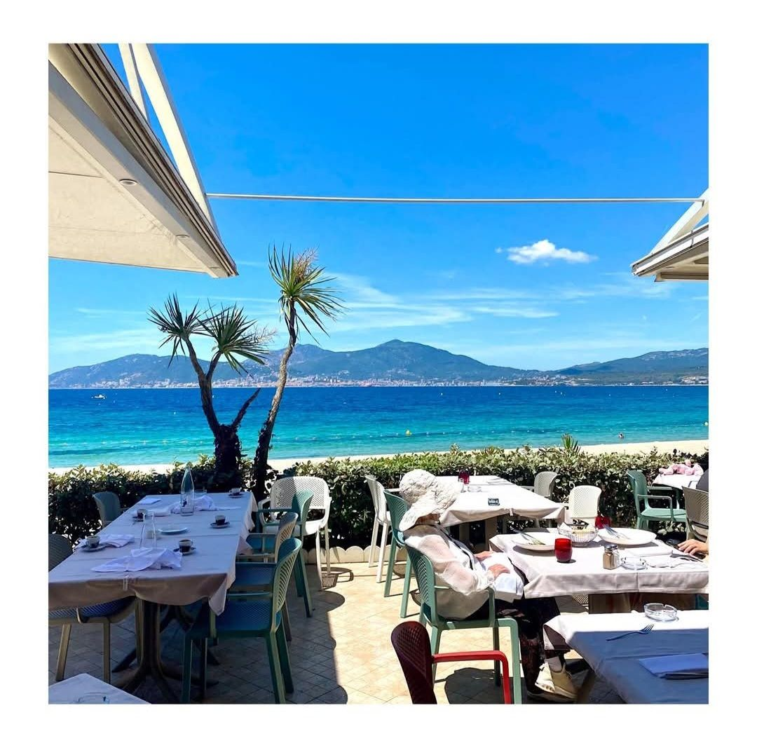 Tables de café en bord de mer sous des auvents blancs, avec une eau turquoise et une montagne au loin sous un ciel bleu.