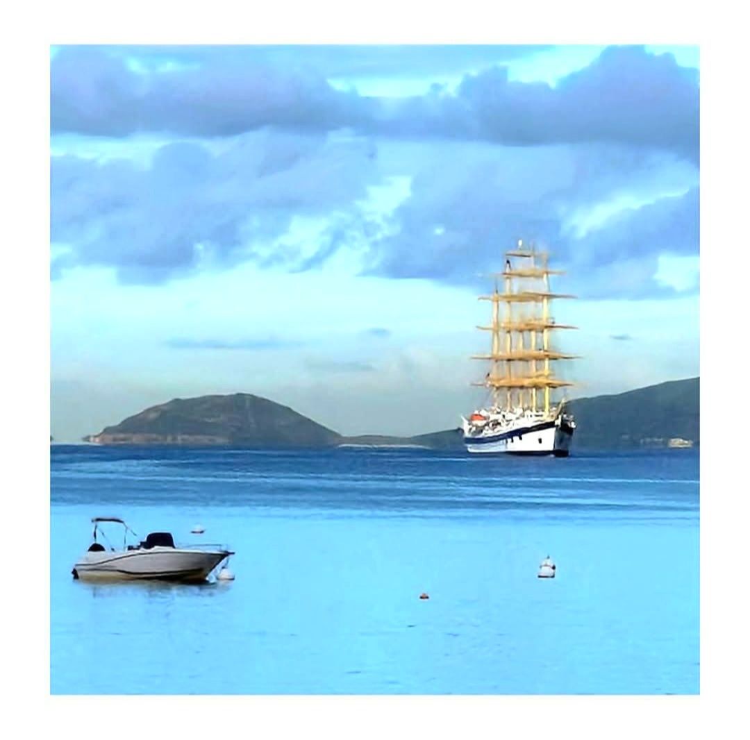 Voilier sur une mer bleue calme avec des îles au loin sous un ciel nuageux, petit bateau à moteur au premier plan.