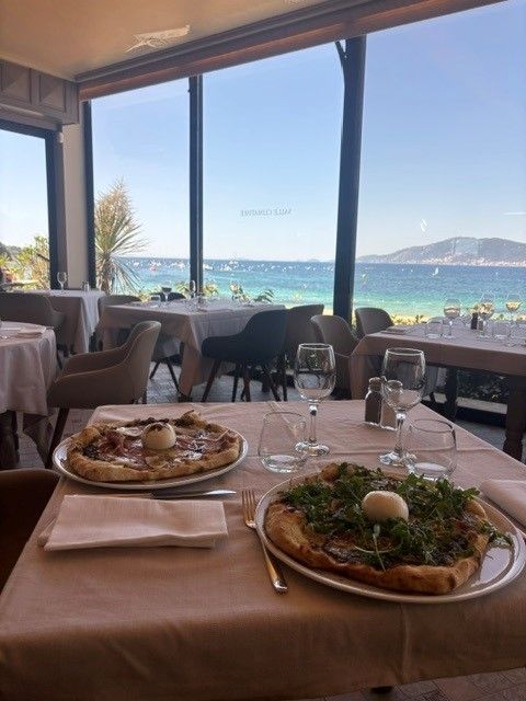 Table de restaurant avec des assiettes de pizza donnant sur une plage ensoleillée et la mer bleue à travers de grandes fenêtres.