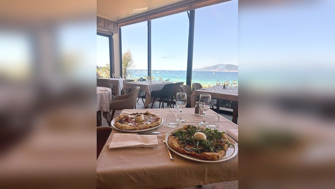 Table de restaurant en bord de mer avec des plats servis à l'assiette et vue sur l'océan à travers les fenêtres ouvertes.