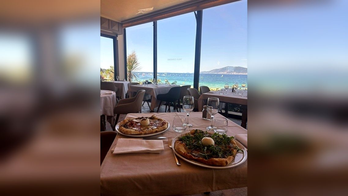 Table à manger près d'une fenêtre donnant sur la mer, avec des assiettes de nourriture, surplombant l'eau bleue et les collines au loin.