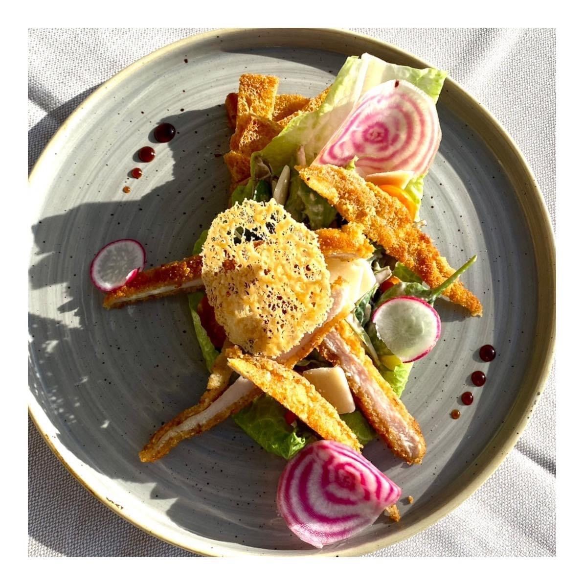 Plat gastronomique composé d'une noix de Saint-Jacques poêlée, de légumes croquants, de volutes violettes et de points de sauce, présenté sur une assiette grise.