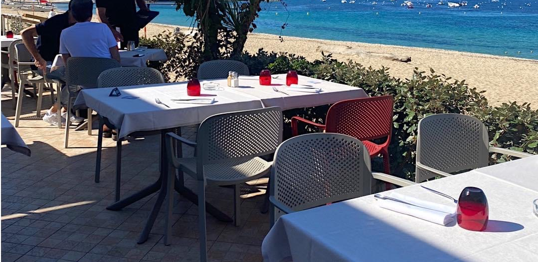 Café en bord de mer avec terrasse, tables et chaises blanches, porte-serviettes rouges et vue sur la mer.