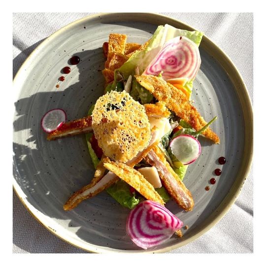 Plat gastronomique composé d'une noix de Saint-Jacques poêlée, de tranches de radis rose, de verdure et de garnitures croustillantes, présenté sur une assiette grise.