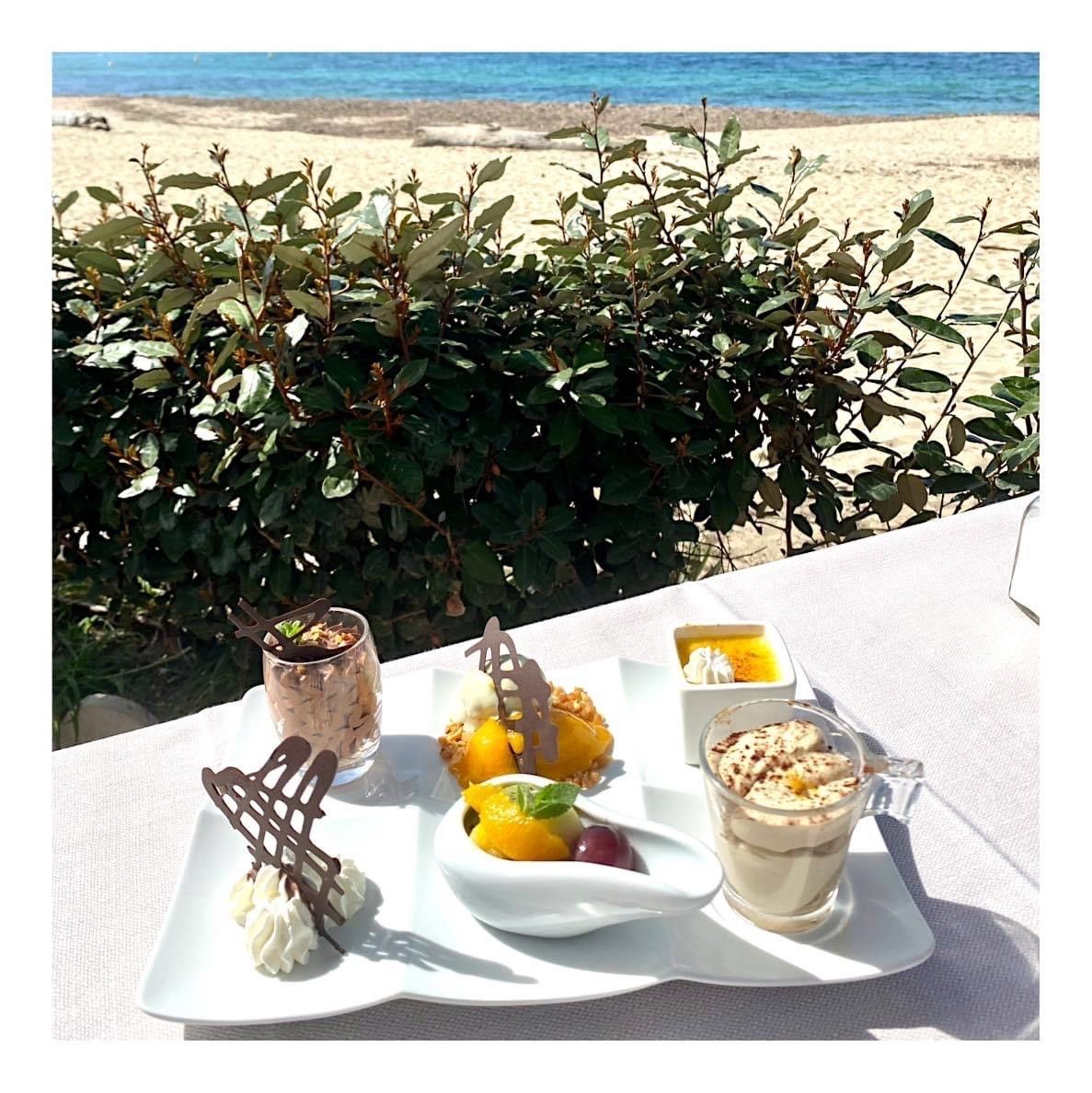 Table en bord de mer avec desserts, fruits et boissons, au milieu des arbustes et des vagues de la mer.