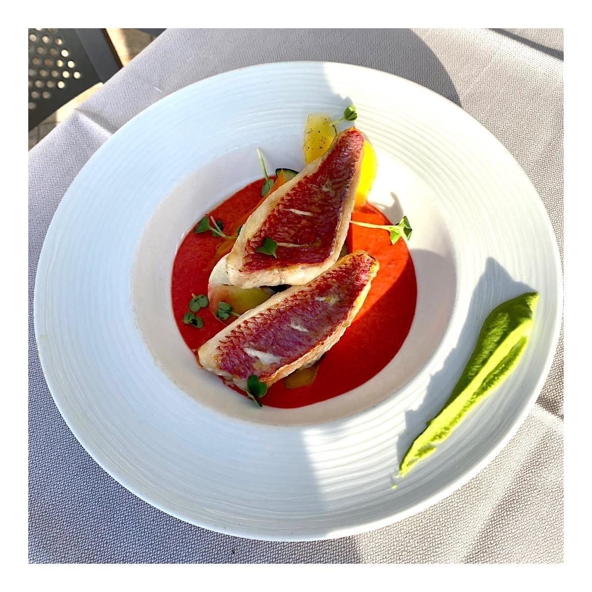 Tranches de figues disposées sur une sauce rouge avec une garniture verte, sur un plat blanc au soleil.