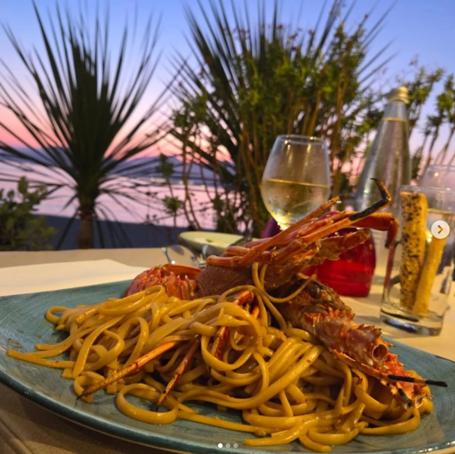 Des pâtes aux crevettes sur une table en bord de mer au coucher du soleil, avec des boissons et des palmiers en arrière-plan.
