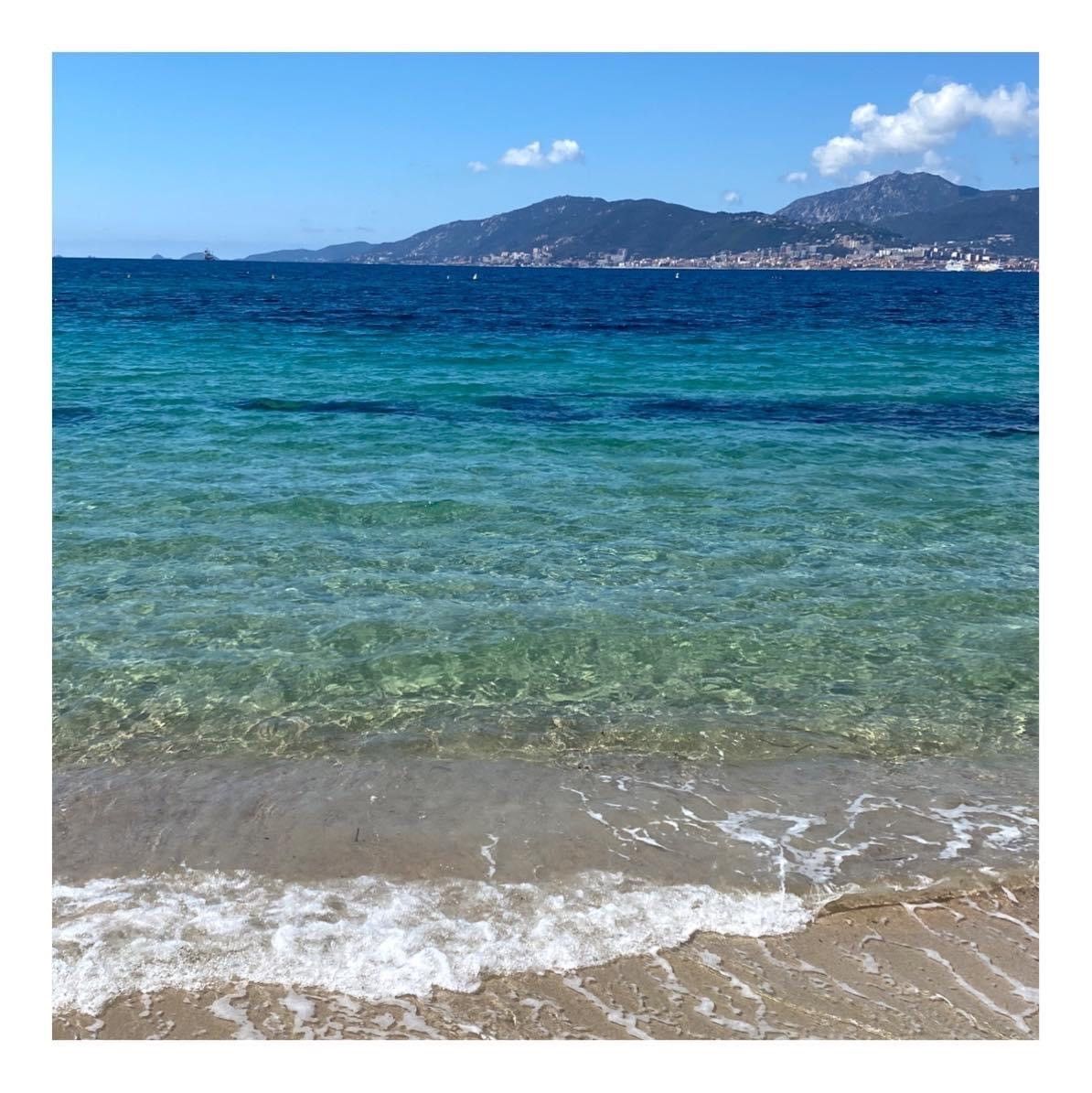 La mer avec de petites vagues et une plage de sable fin, avec des montagnes à l'horizon sous un ciel lumineux