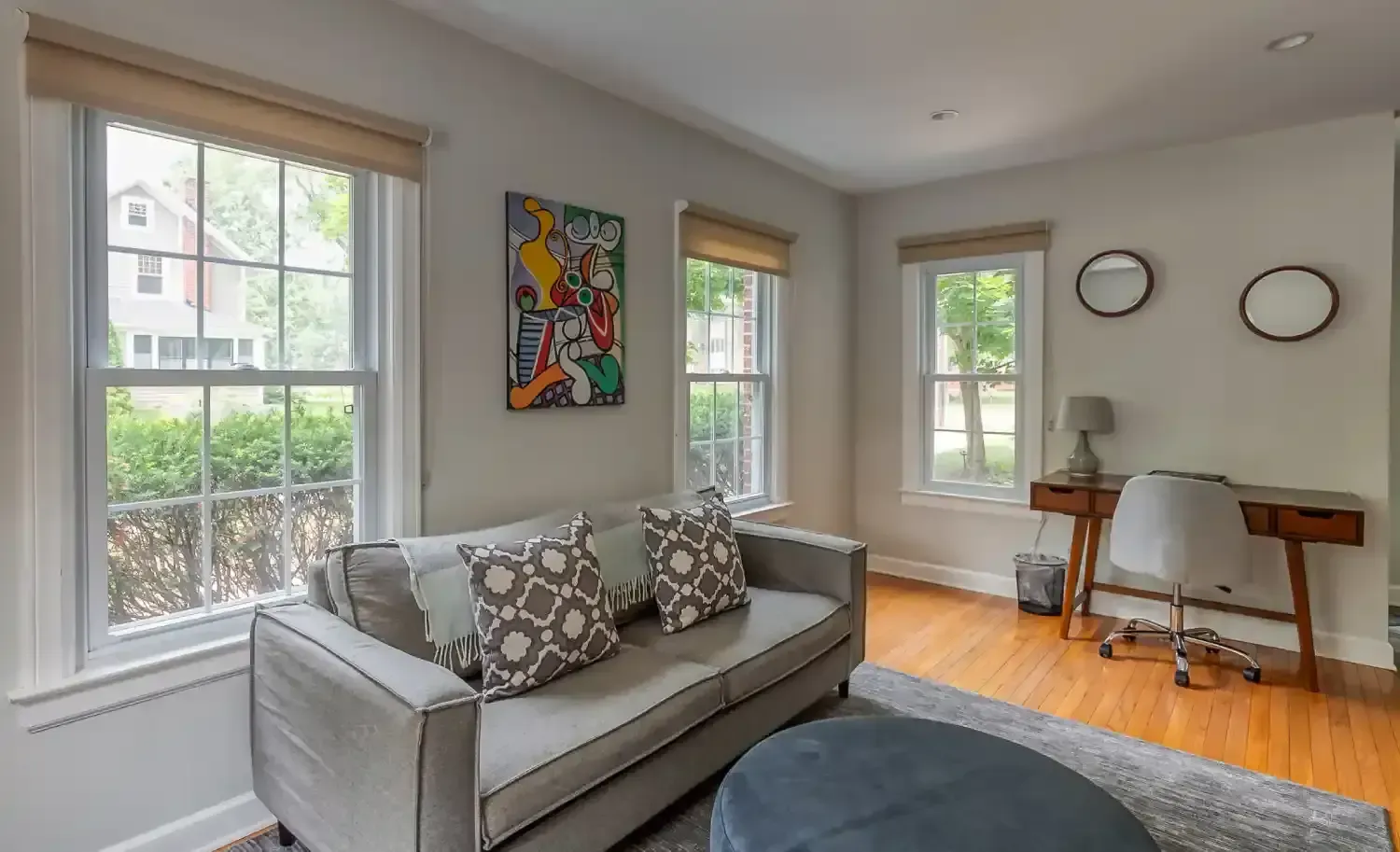 Living room with sofa, desk, artwork, and three windows, light wood floors.