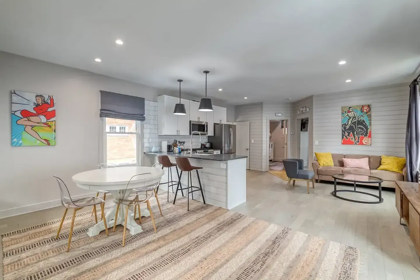 Open-concept living space with kitchen, dining, and seating areas; white walls, light wood floors, art, and rug.