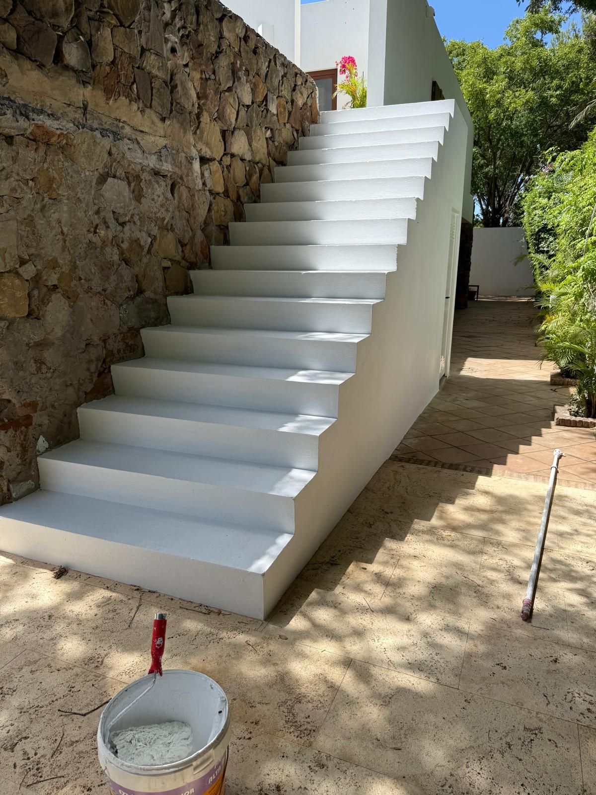 Escalera blanca exterior en construcción junto a un muro de piedra, con un cubo de yeso en primer plano.