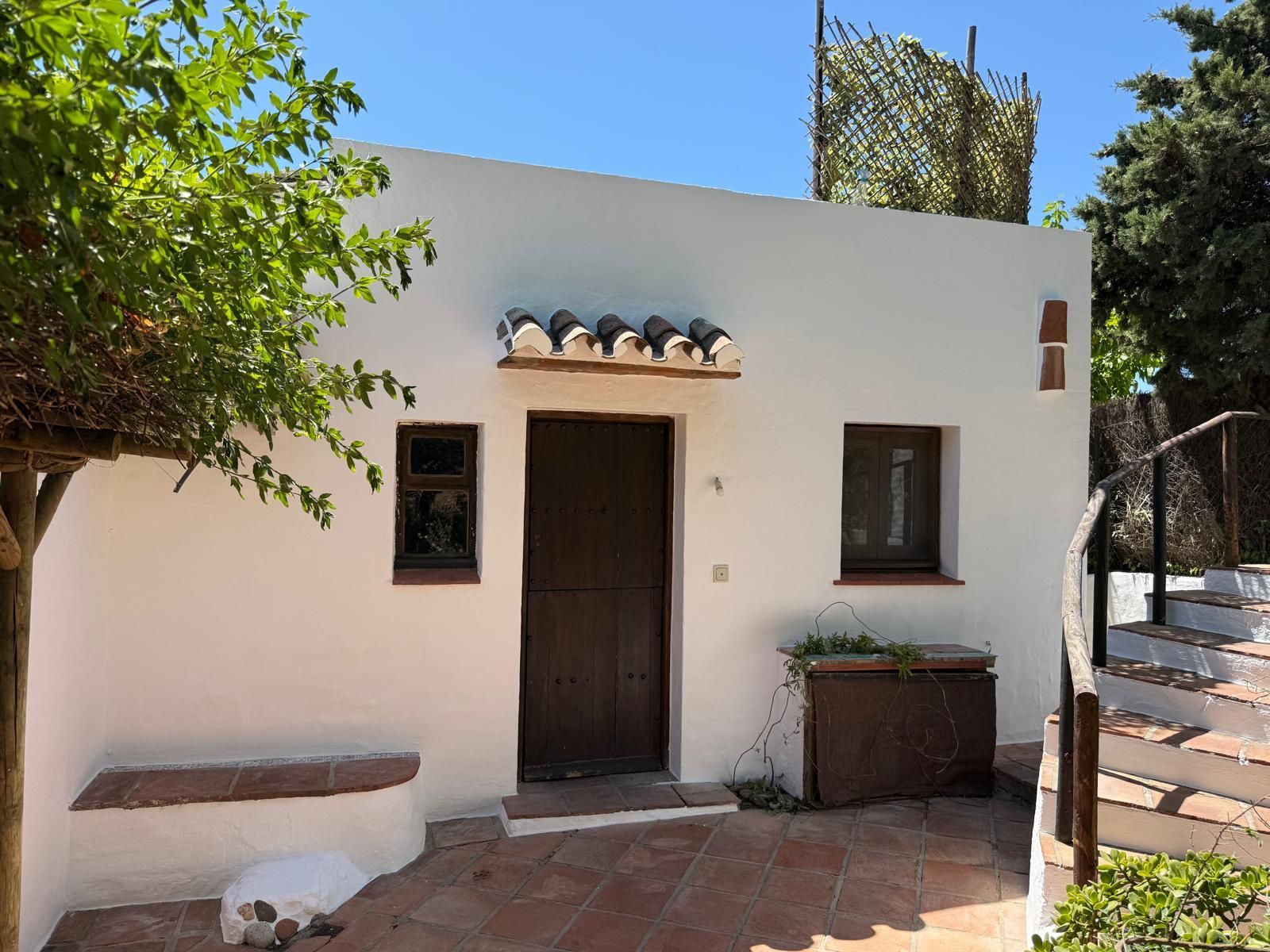 Pequeña casa blanca de estuco con puerta de madera oscura, dos ventanas y un banco en un patio soleado.