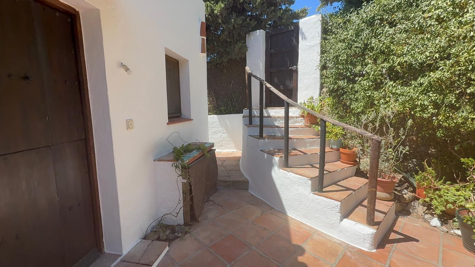 Patio soleado con escaleras, barandilla negra, baldosas de terracota, paredes blancas y plantas en macetas.