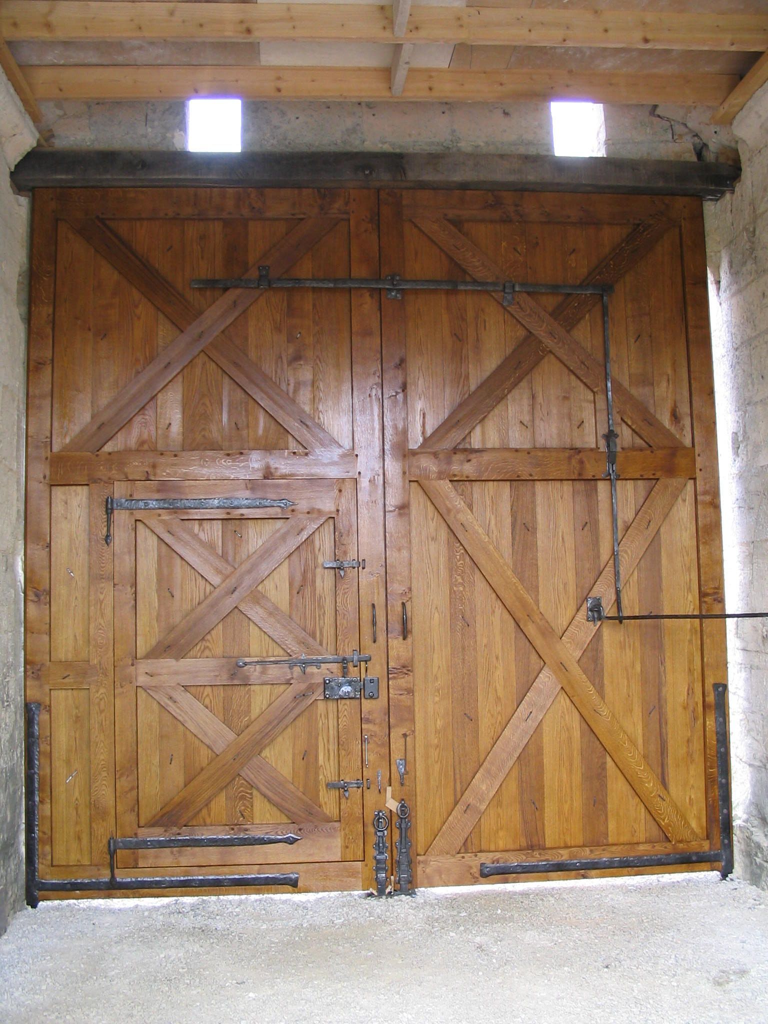 Double porte en bois avec ferrures métalliques, encastrée dans un mur en pierre, deux petites fenêtres au-dessus.
