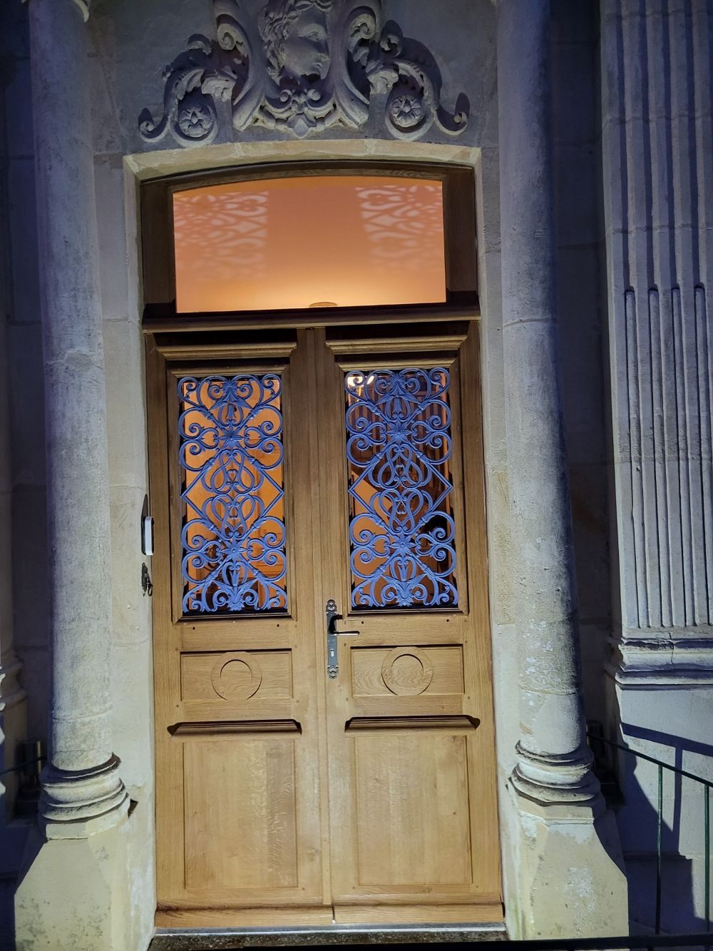 Double porte en bois ornée de ferronneries, surmontée d'une fenêtre et de sculptures décoratives, flanquée de piliers.