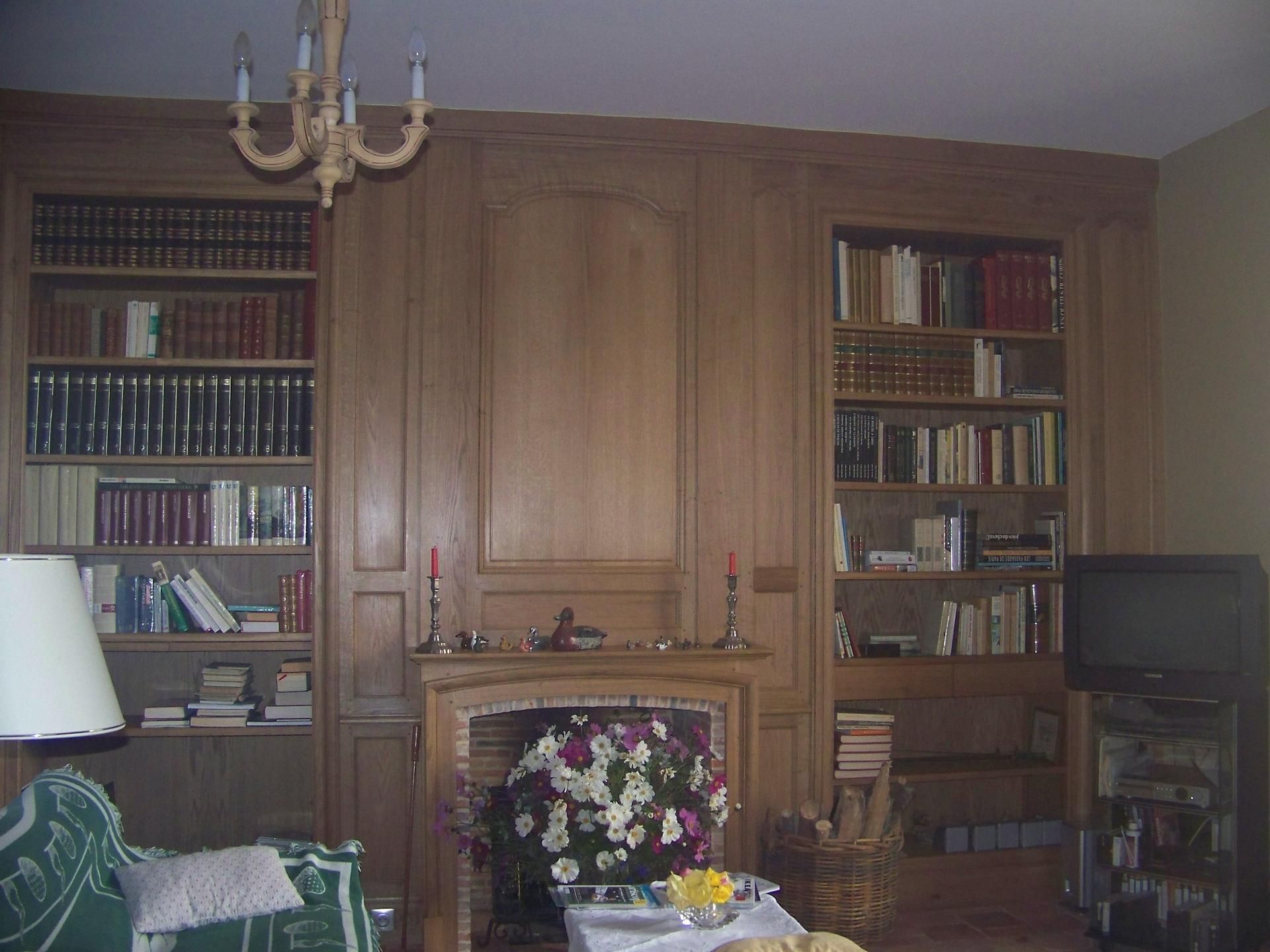 Pièce lambrissée avec des bibliothèques de part et d'autre d'une cheminée et d'un lustre.