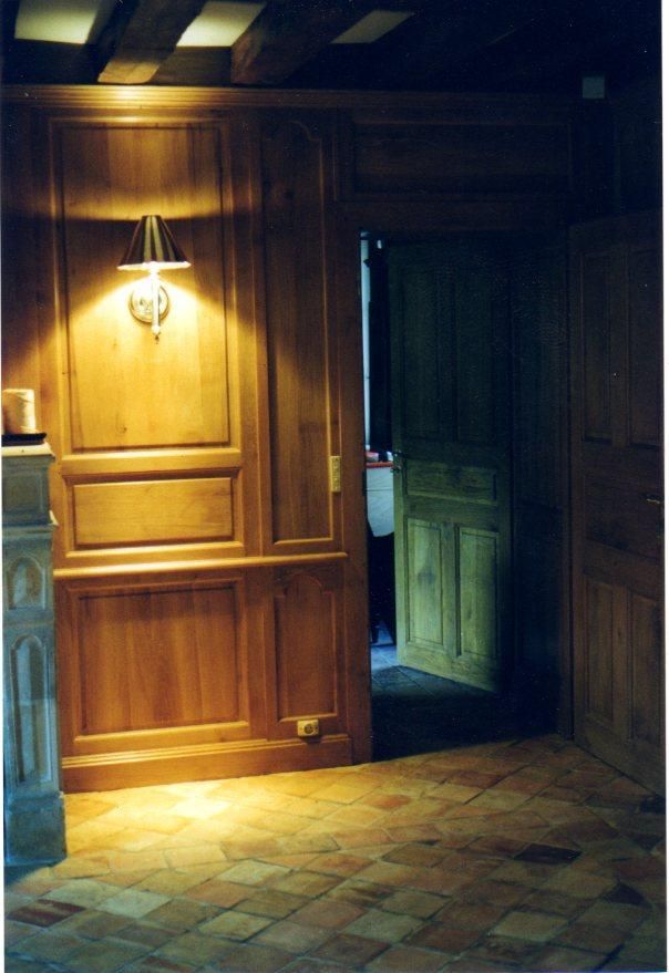 Mur lambrissé avec une applique murale éclairée à côté d'une porte ouverte. Sol carrelé de rouge.