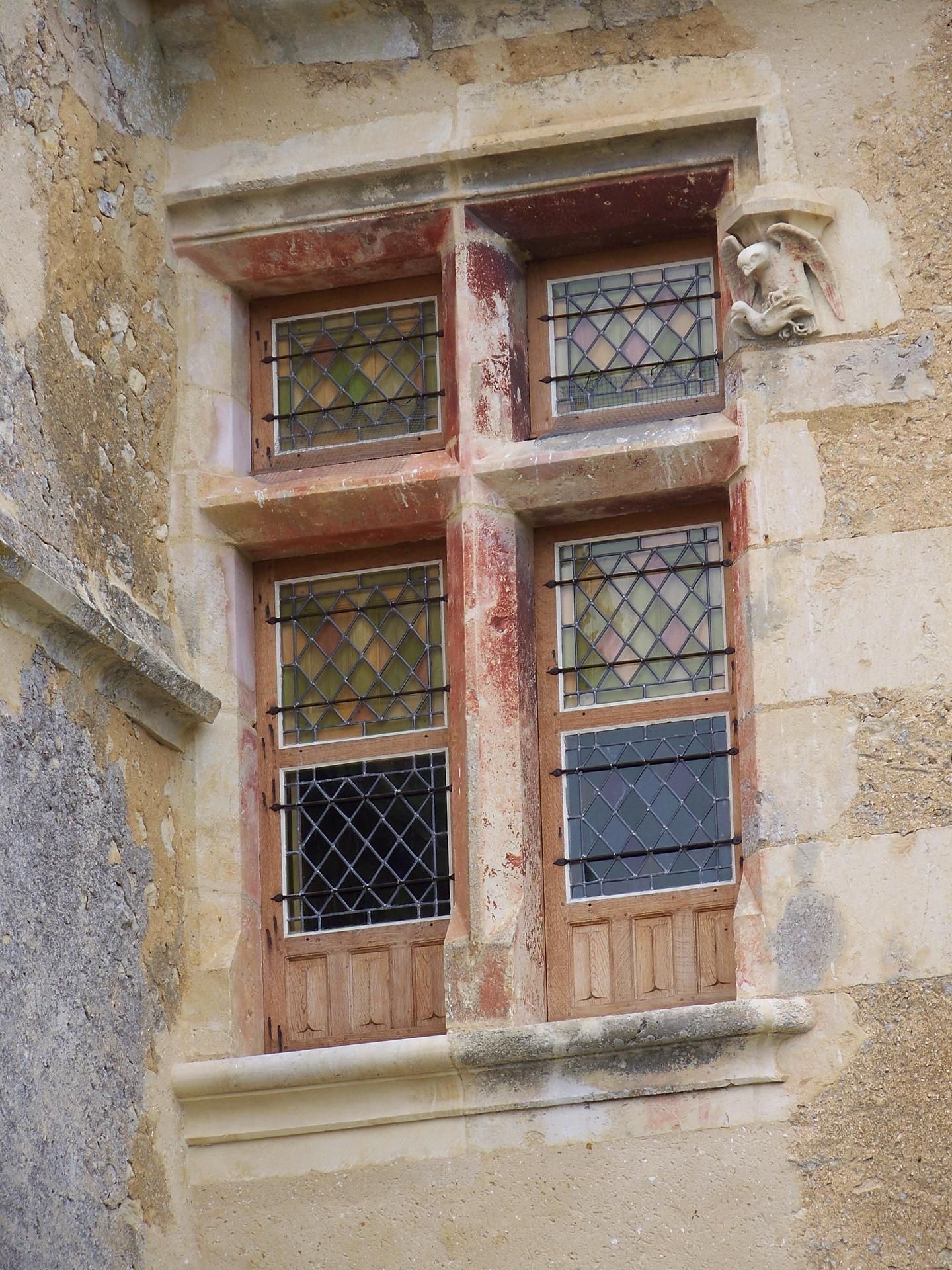 Fenêtre en pierre à quatre carreaux, cadres en bois, grilles en métal noir et une sculpture en pierre ornementale sur la droite.
