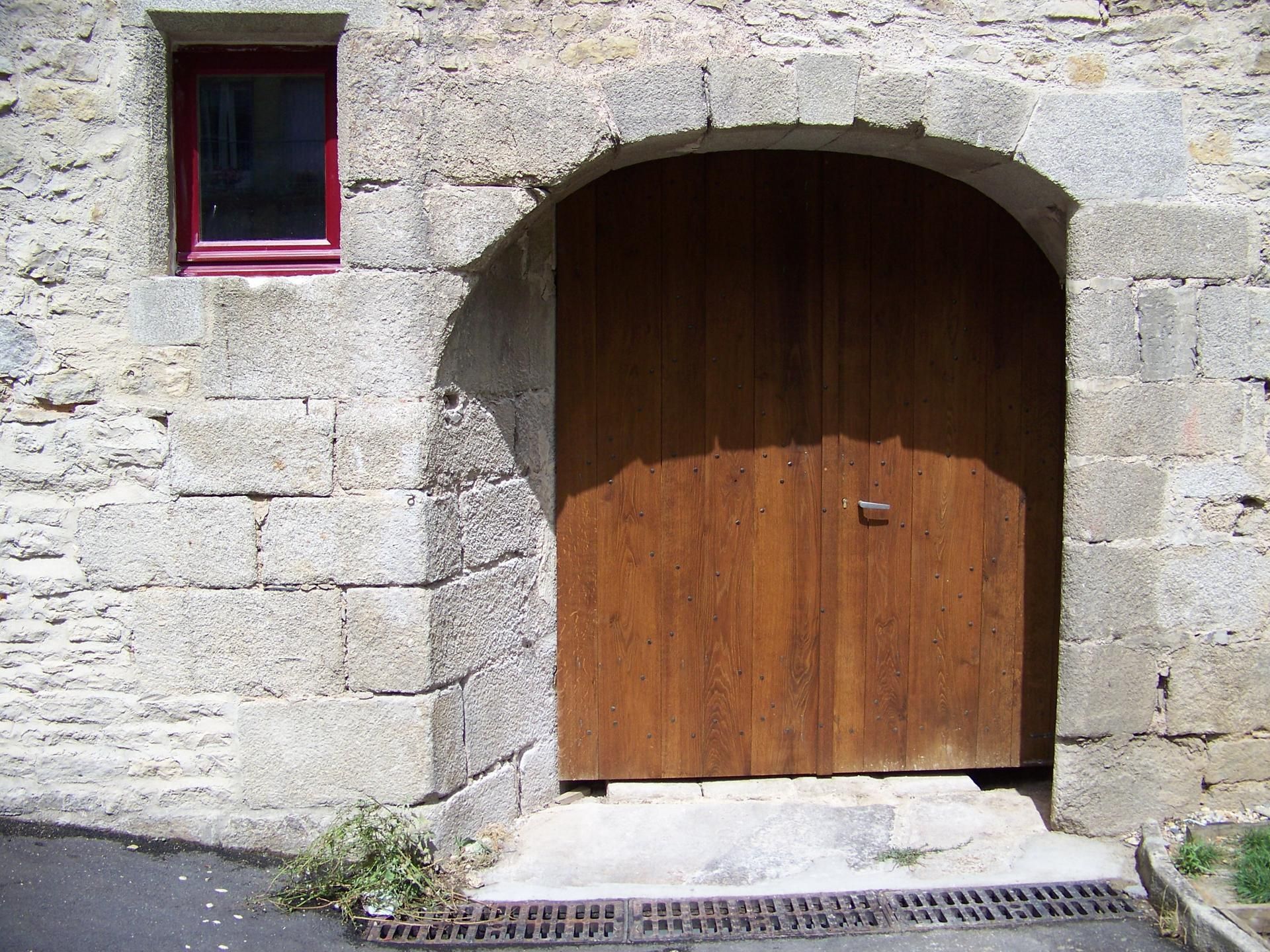 Bâtiment en pierre avec une porte cintrée en bois et une petite fenêtre à cadre rouge.