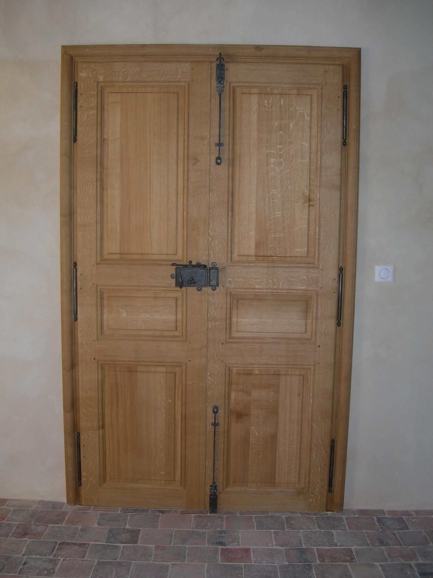 Double porte en bois avec ferrure en fer, encastrée dans un mur clair, sur un sol en briques.