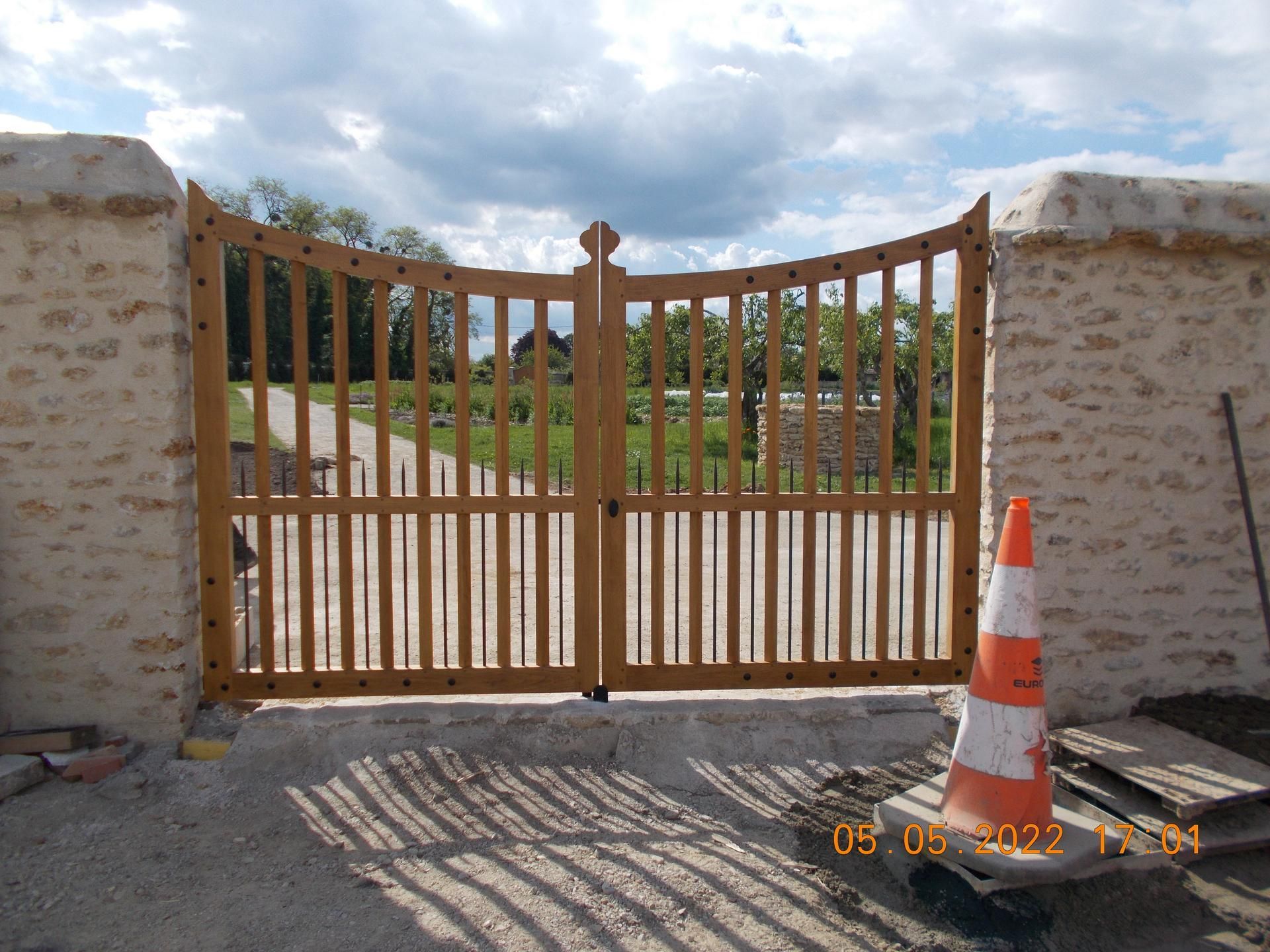 Portes en bois dans un mur de pierre, cône de signalisation orange au premier plan, ciel nuageux en arrière-plan.
