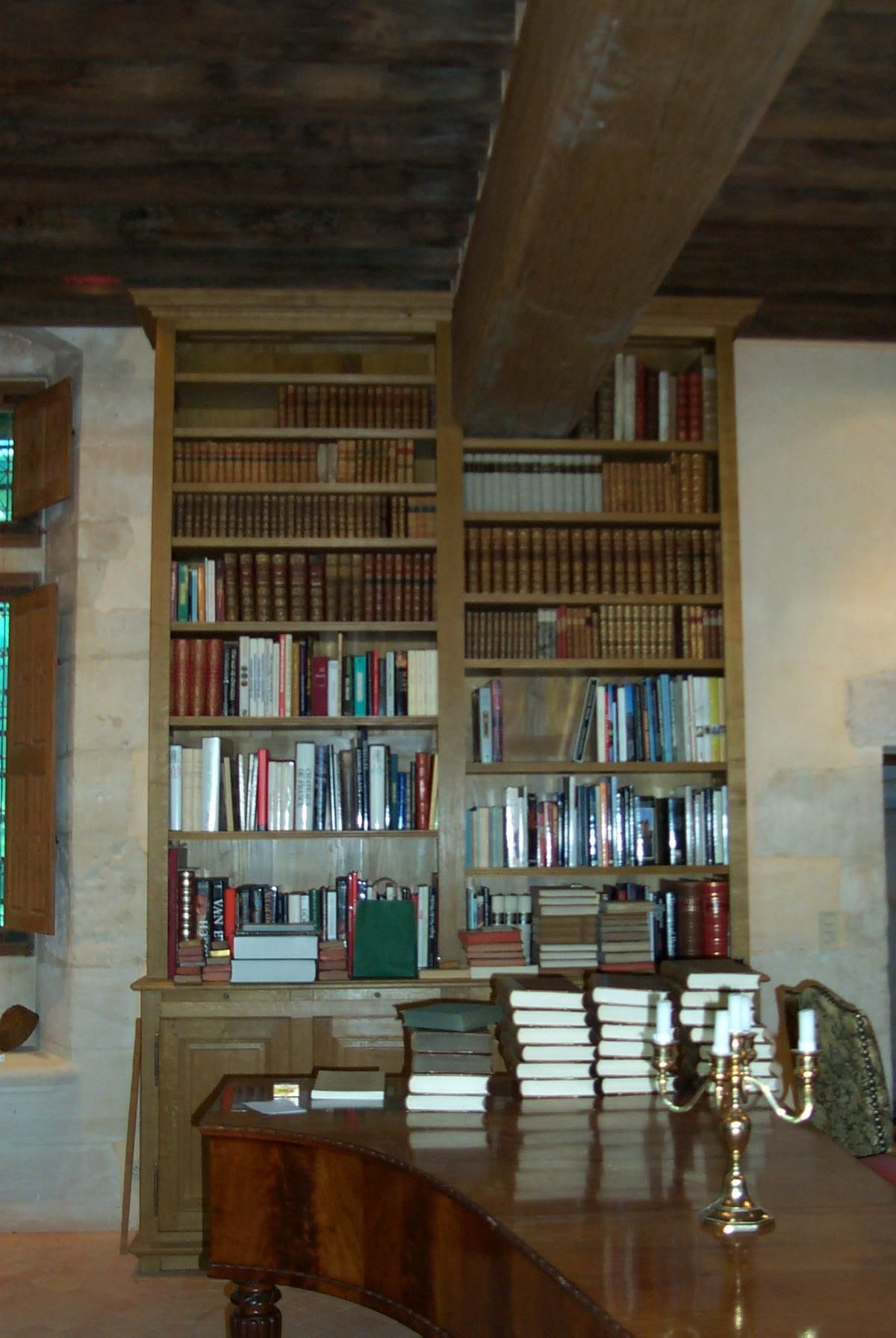 Des étagères en bois remplies de livres sont adossées à un mur. Une poutre en bois est suspendue au-dessus. Une table est placée devant.