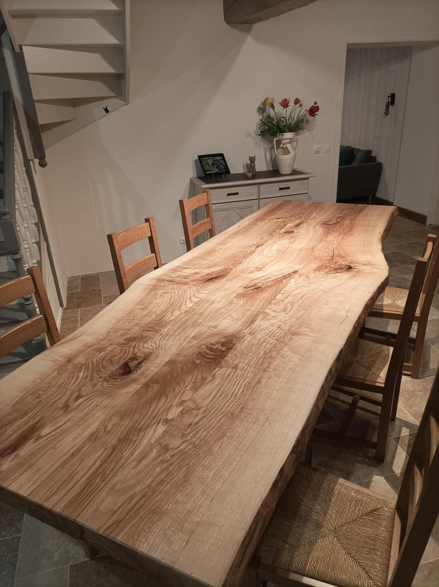 Table à manger en bois avec chaises dans une pièce rustique ; fleurs et armoire en arrière-plan.