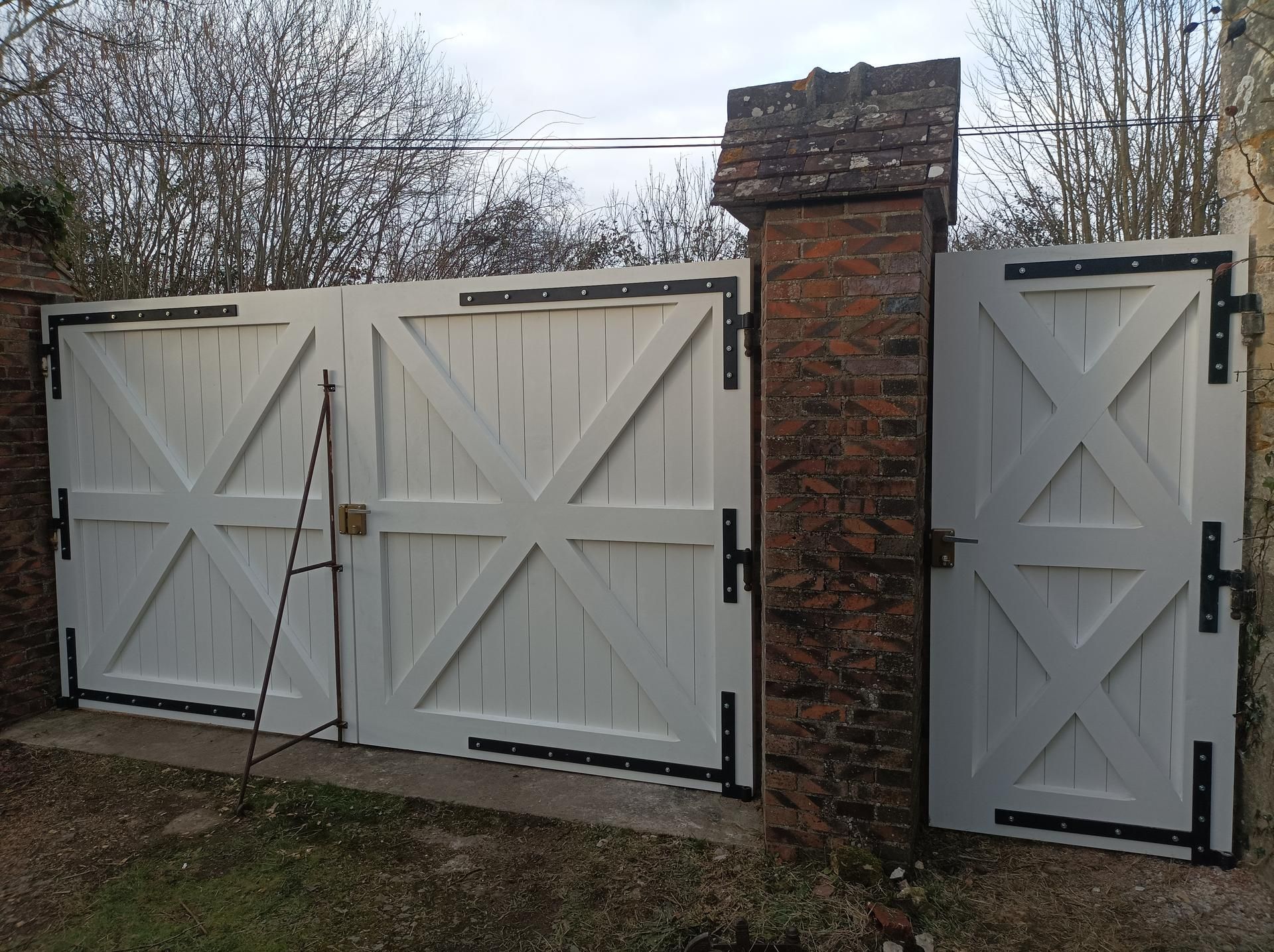 Des portails en bois blanc, avec des charnières noires et des entretoises en forme de X, sont fixés à des piliers en brique.